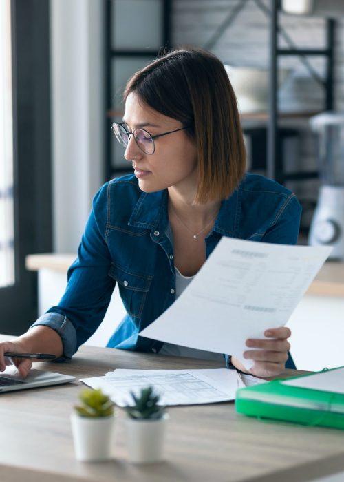 young woman working with computer while consulting some invoices and documents in the kitchen